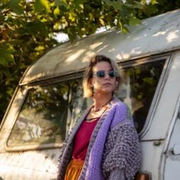 Portret kobiety w okularach przeciwsłonecznych, opierającej się o starą, białą przyczepę kempingową w plenerze, z liśćmi drzew w tle.