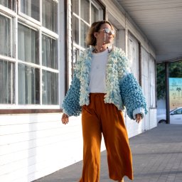 Justyna Jeż - Kobieta w puszystym, błękitnym kardiganie i szerokich, pomarańczowych spodniach spaceruje na tle białej ściany z oknami. Uwagę przykuwają okulary na sznurku z koralików.