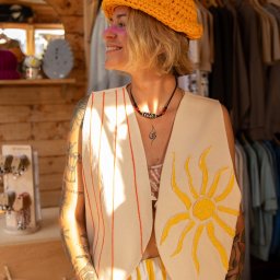Justyna Jeż - Kobieta z różowym makijażem pod okiem, w żółtym kapeluszu z włóczki i kamizelce ze słońcem, w sklepie z odzieżą na wieszakach i drewnianymi półkami.