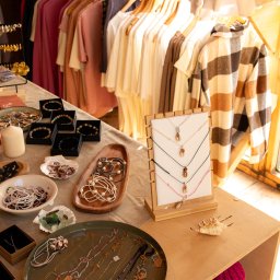 Justyna Jeż - Stoisko z biżuterią i odzieżą: naszyjniki z zawieszkami, bransoletki w pudełkach, ubrania na wieszakach. Jasne, naturalne światło podkreśla detale rękodzieła.