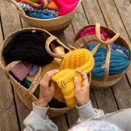 Justyna Jeż - Ręce trzymają żółtą, dzierganą czapkę z drewnianym logo na tle koszy z kolorowymi czapkami na drewnianym podłożu. Widok z góry.
