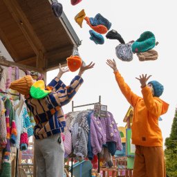 Justyna Jeż - Dwie osoby w kolorowych ubraniach podrzucają w górę ręcznie robione czapki na tle wieszaków z odzieżą i placu zabaw, tworząc dynamiczne ujęcie.