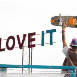 Justyna Jeż - Osoba z tatuażami unosi deskorolkę na tle napisu 'LOVE IT', widok z tyłu, w tle jasne niebo i elementy infrastruktury miejskiej.