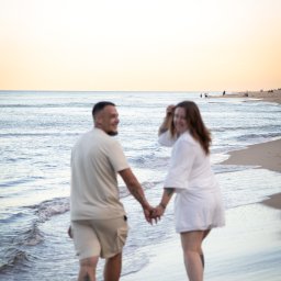 Justyna Jeż - Para trzymająca się za ręce spaceruje brzegiem morza o zachodzie słońca. Kobieta odwraca się z uśmiechem. Widoczne tatuaże na ciele mężczyzny.