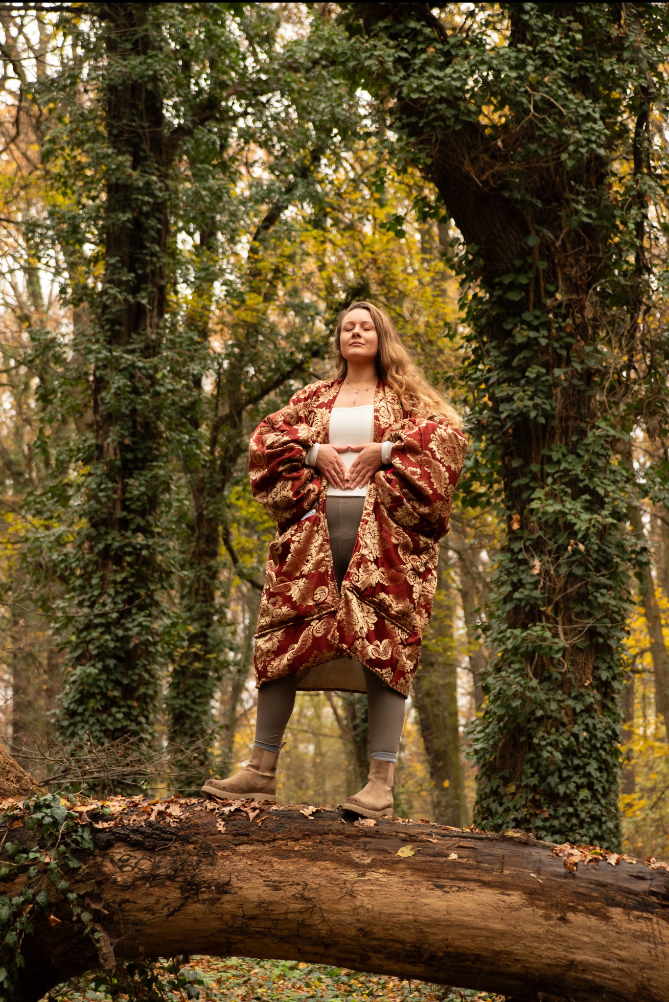 Kobieta w kwiecistym szlafroku stoi na powalonym pniu w lesie, z zamkniętymi oczami i dłońmi ułożonymi w kształt serca na brzuchu. Jesienne drzewa w tle.