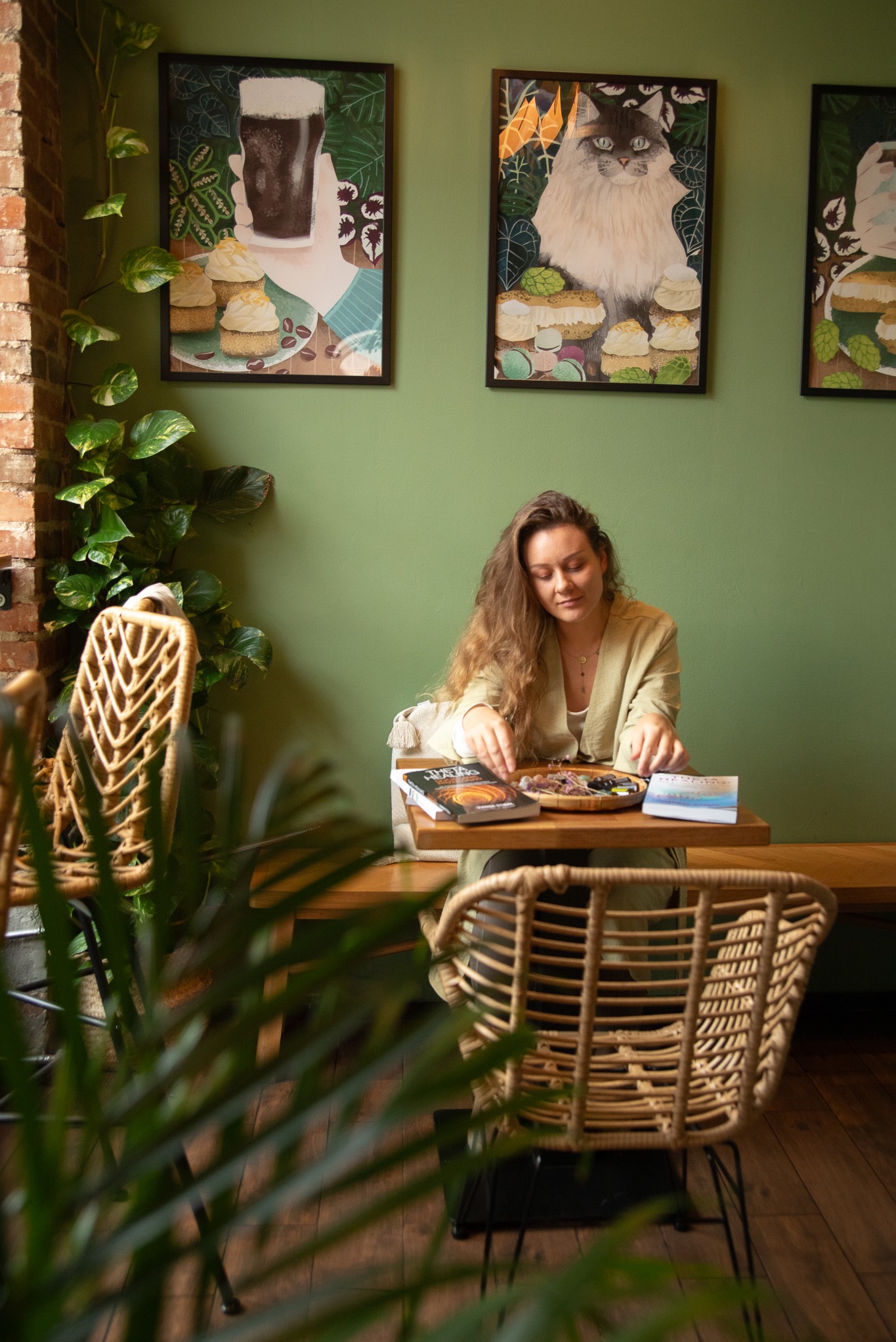 Kobieta siedzi przy drewnianym stole w kawiarni z zielonymi ścianami, układając kamienie na drewnianej tacy. Na ścianie wiszą grafiki z motywami jedzenia i zwierząt.