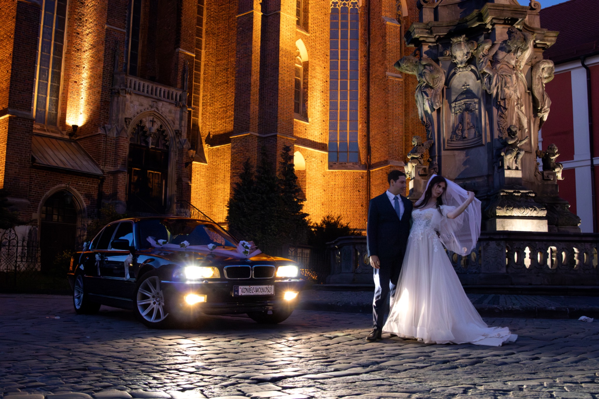 Para młoda w sukni ślubnej i garniturze pozuje przy samochodzie z dekoracjami na tle oświetlonej, gotyckiej architektury miejskiej.