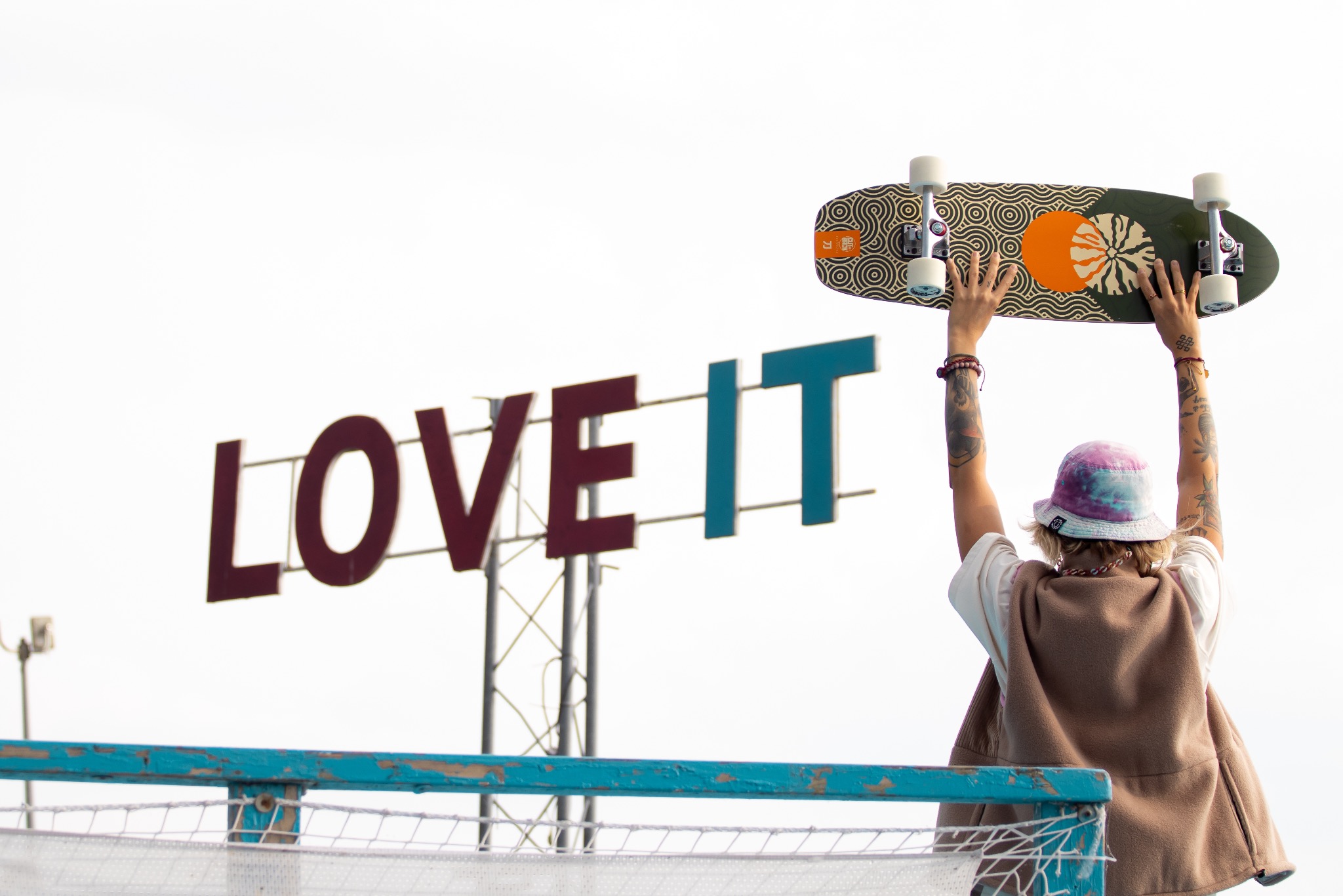 Osoba z tatuażami unosi deskorolkę na tle napisu 'LOVE IT', widok z tyłu, w tle jasne niebo i elementy infrastruktury miejskiej.