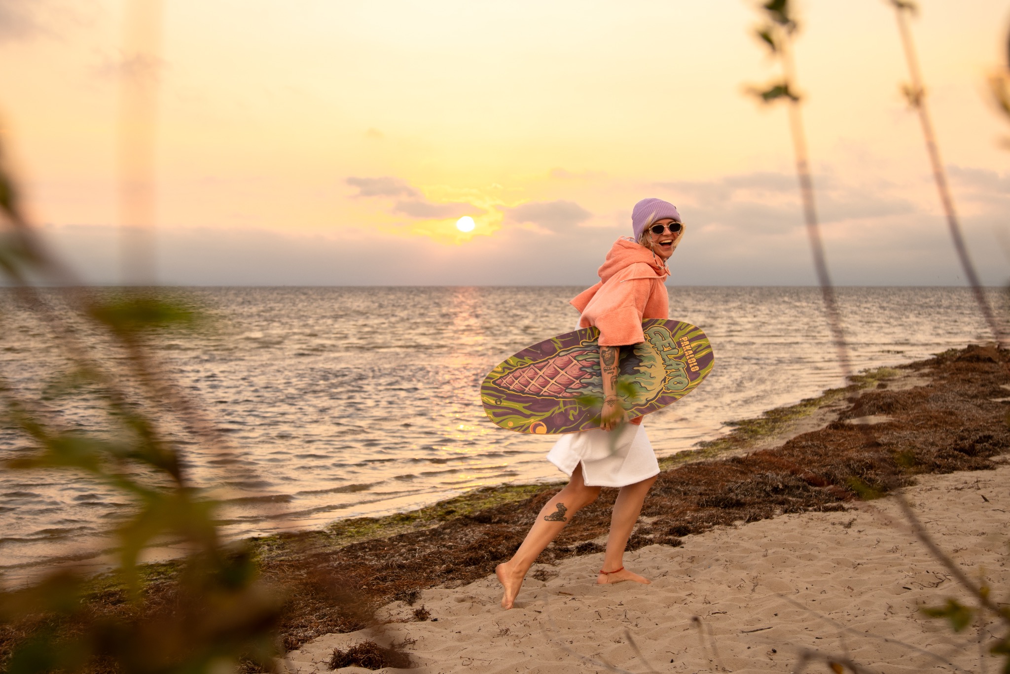 Uśmiechnięta kobieta w okularach i czapce niesie deskę na plaży o zachodzie słońca. Widoczne morze i piasek, a w tle zachodzące słońce.