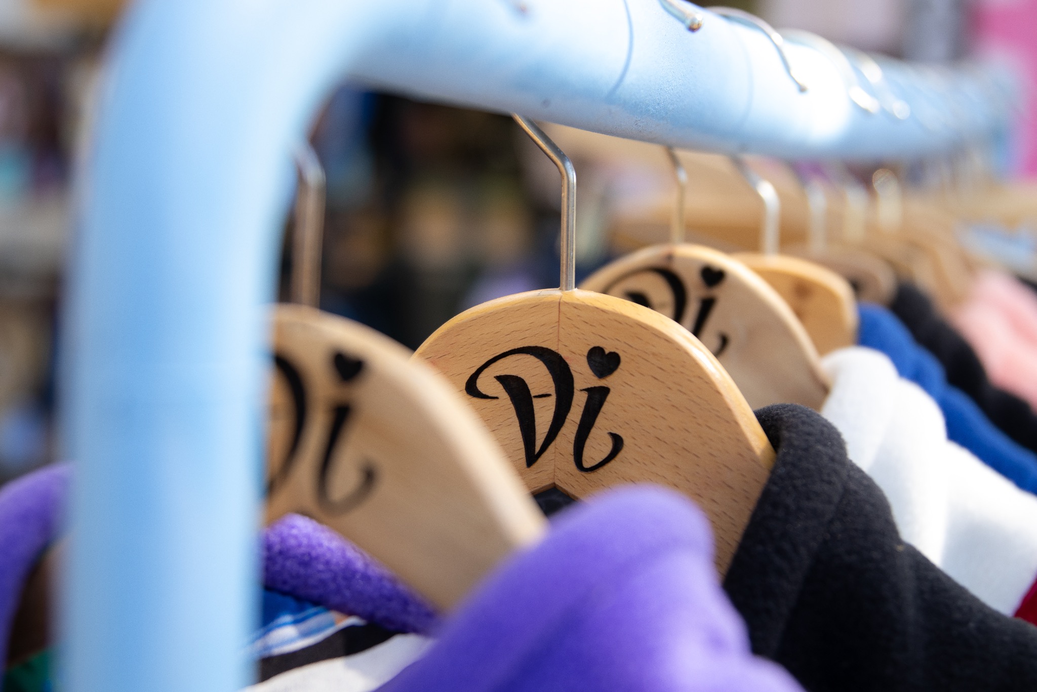 Rząd drewnianych wieszaków z wygrawerowanym logo 'Vi' w kształcie serca, na których wiszą kolorowe bluzy. Lekkie rozmycie tła, błękitny metalowy stelaż.