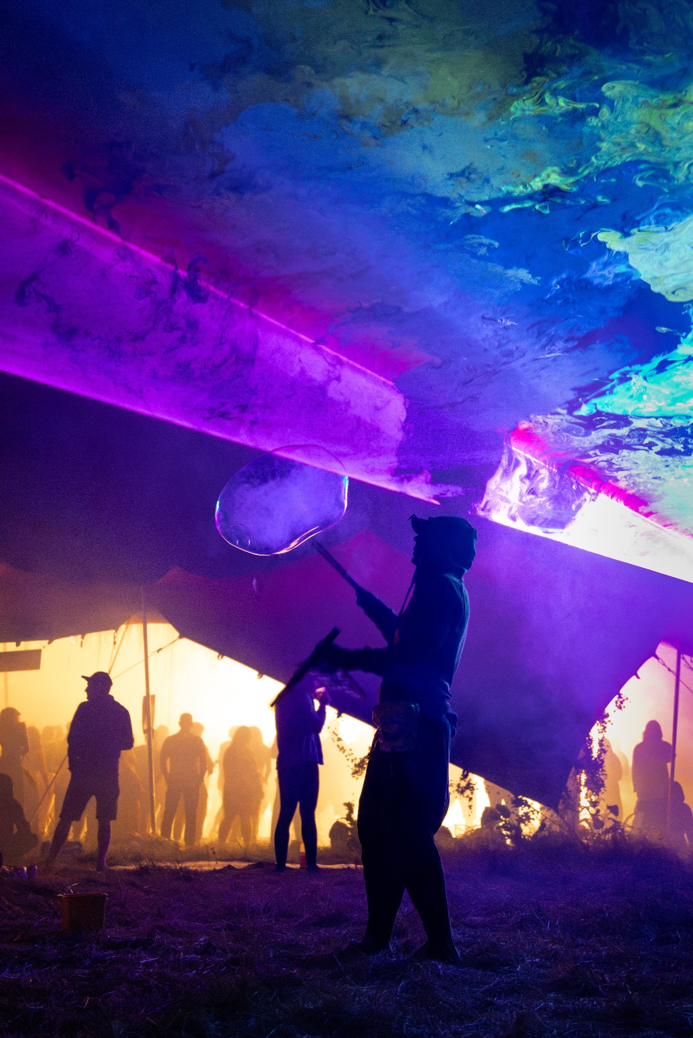 Sylwetka osoby puszczającej bańki mydlane na tle sceny z laserami i tłumu podczas eventu. Dominują fioletowe i żółte barwy, tworząc unikalny efekt wizualny.