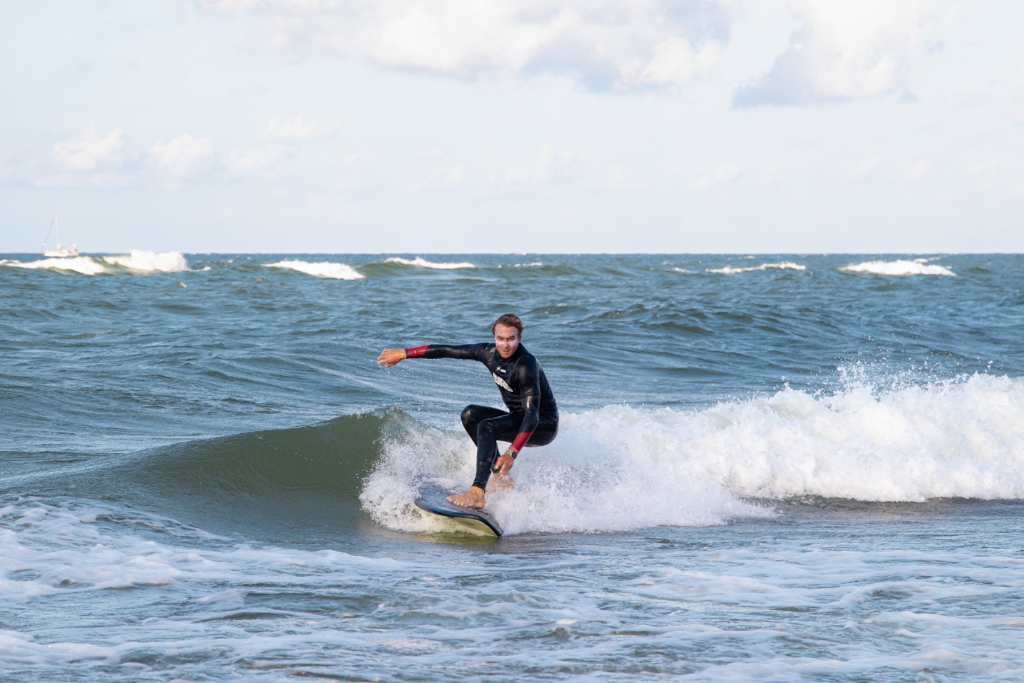 Surfer w czarnym kombinezonie na desce, dynamicznie pokonuje falę. W tle błękitne morze z delikatnymi falami i żaglówka. Jasne niebo z chmurami.