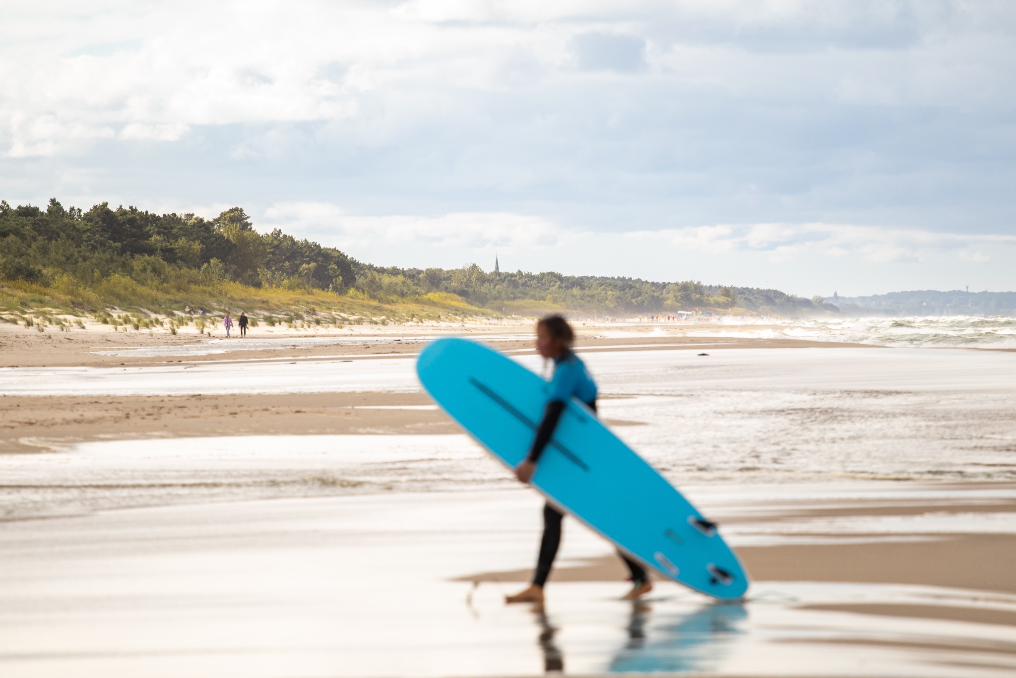 Rozmyta sylwetka surfera z błękitną deską na plaży, w tle zalesione wydmy i morze z falami. Jasne, pochmurne niebo nadaje scenie neutralny, naturalny charakter.