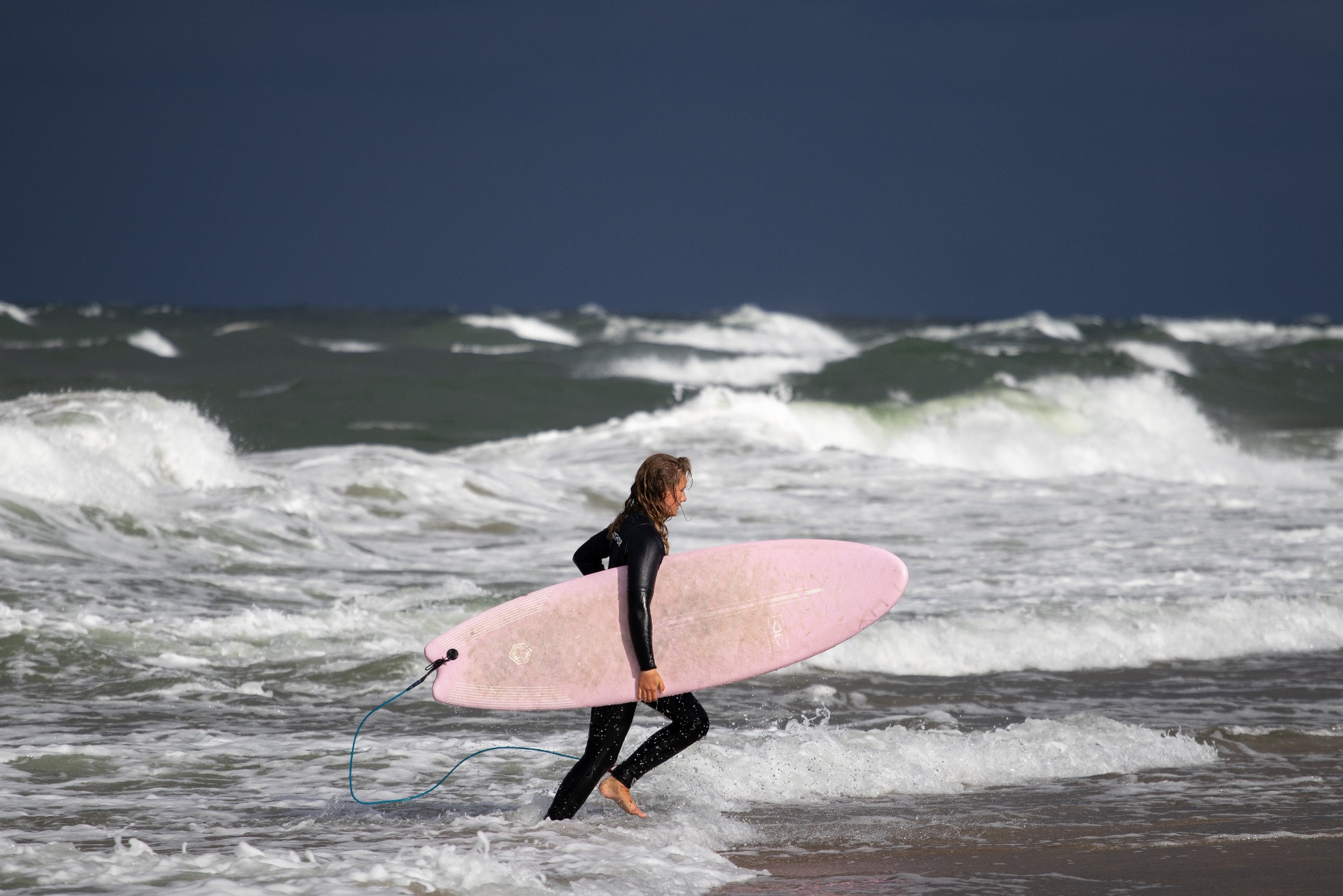 Surferka w czarnym kombinezonie biegnie przez fale z różową deską surfingową na tle wzburzonego morza i ciemnego nieba. Dynamiczne ujęcie z bliska.