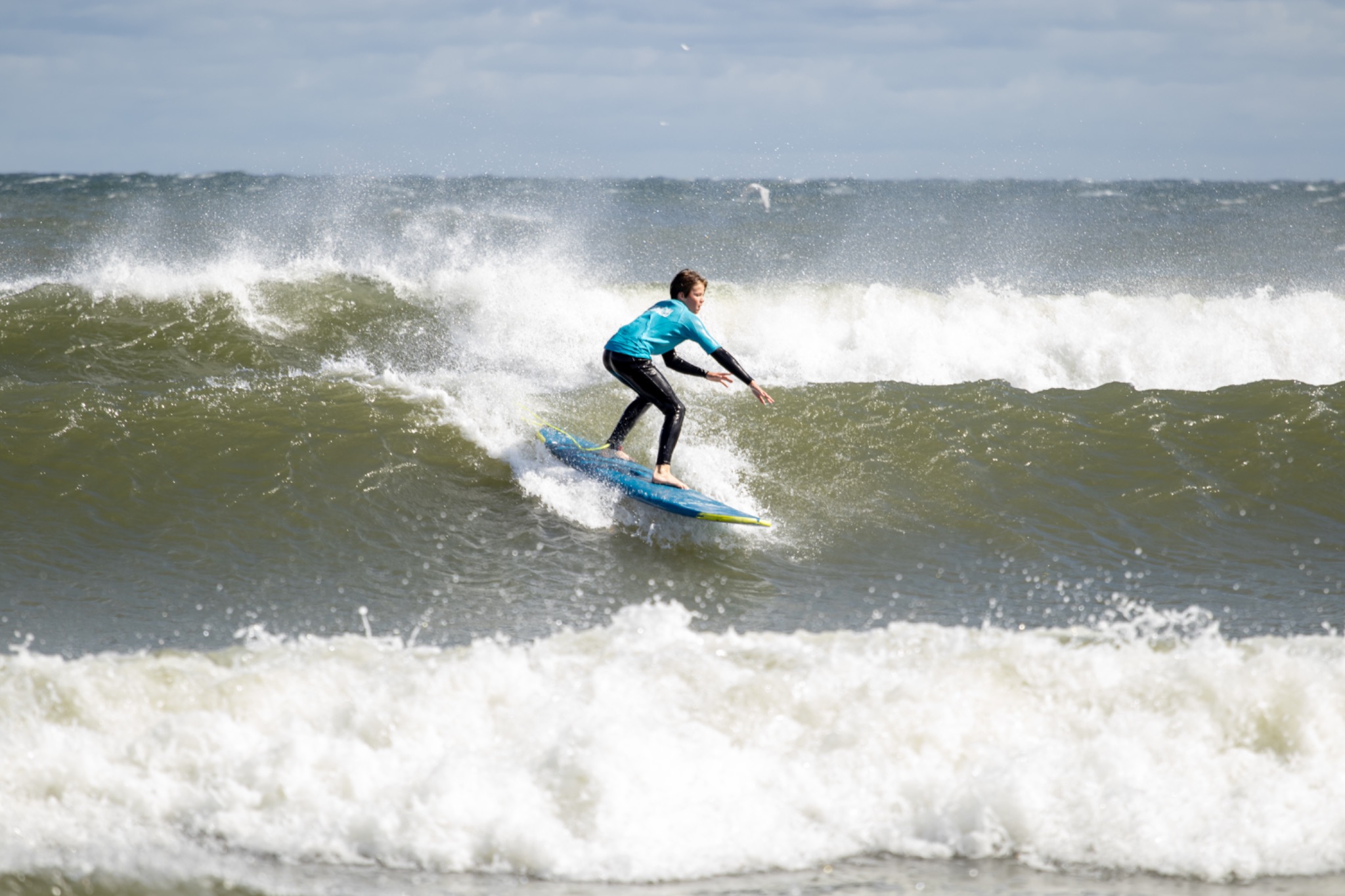 Surfer w turkusowej koszulce płynie na błękitnej desce po wzburzonym morzu. W tle biała piana fal i pochmurne niebo. Dynamiczne ujęcie z bliska.