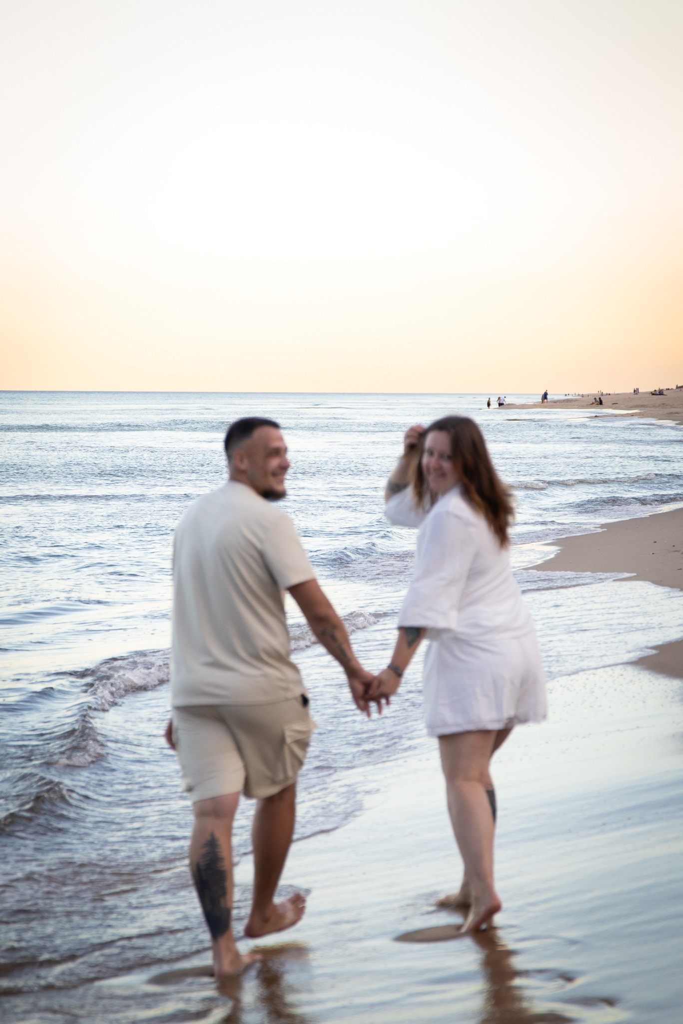 Para trzymająca się za ręce spaceruje brzegiem morza o zachodzie słońca. Kobieta odwraca się z uśmiechem. Widoczne tatuaże na ciele mężczyzny.