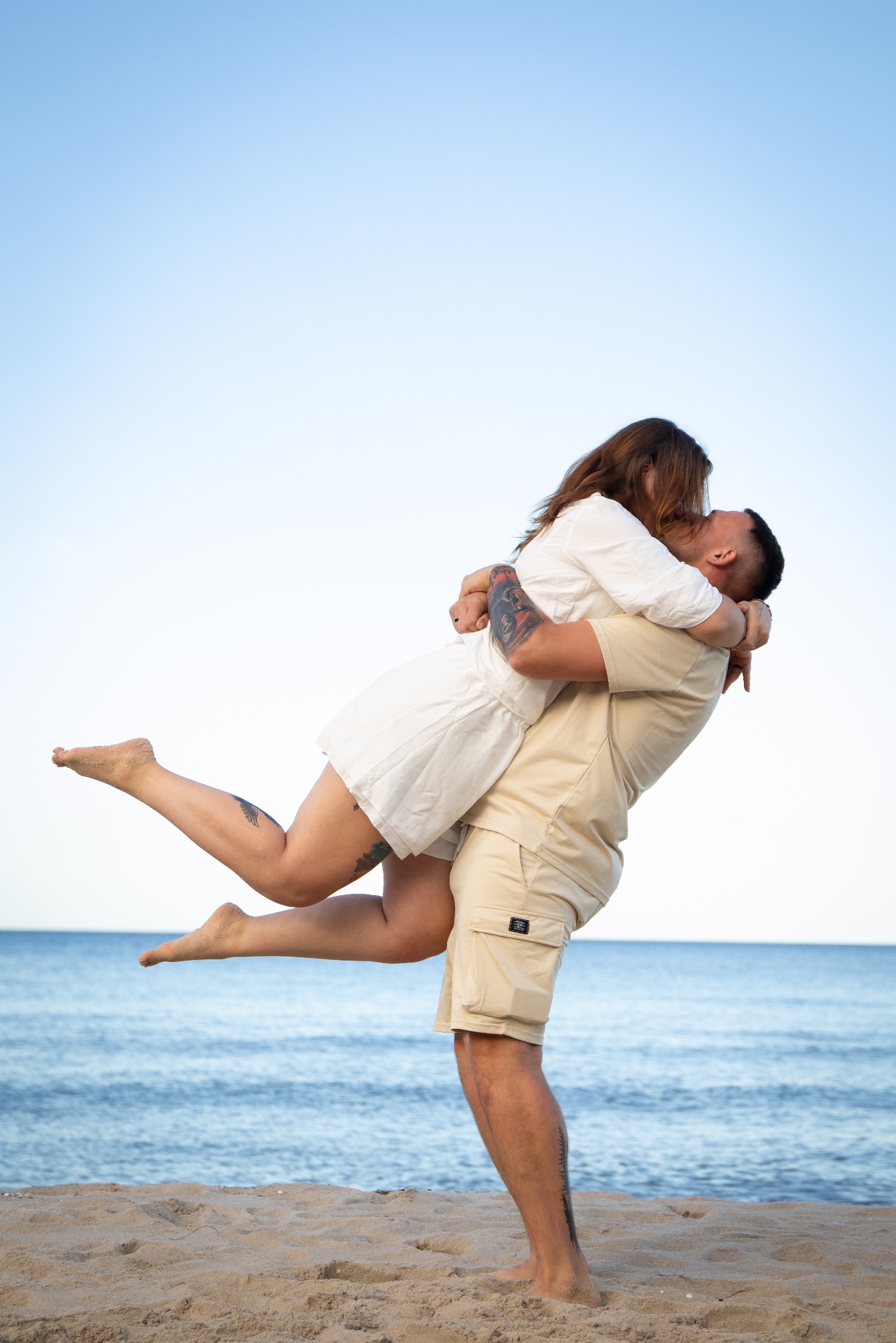 Para zakochanych na plaży we Wrocławiu. Mężczyzna podnosi kobietę, obejmując ją w talii. Oboje ubrani na jasno, w tle morze i błękitne niebo. Widoczne tatuaże na ciele.