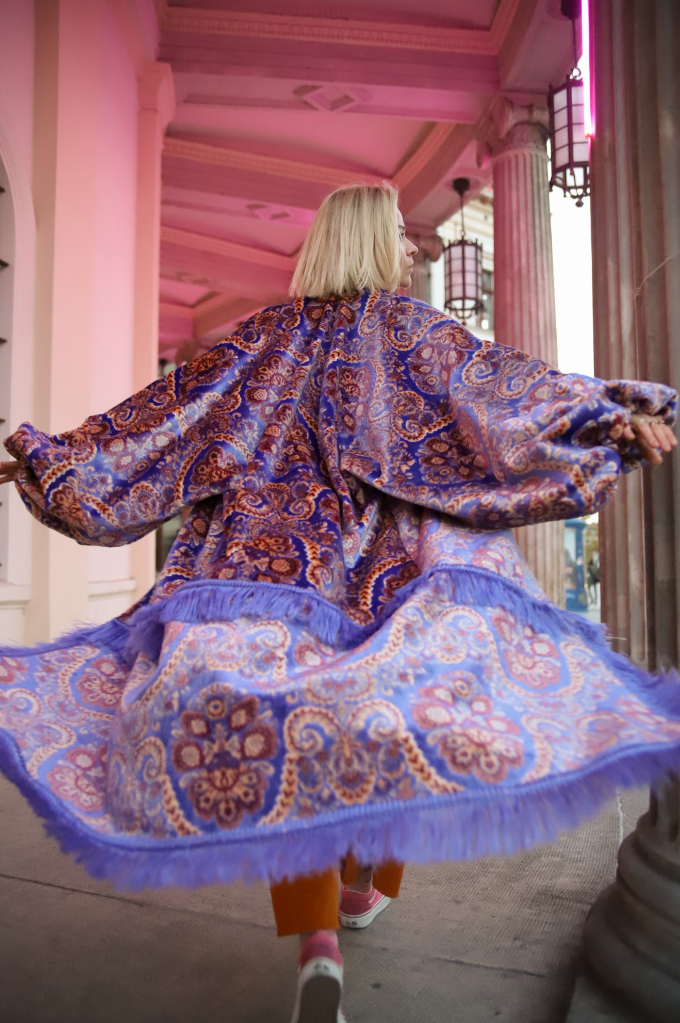 Kobieta w bogato zdobionej, fioletowo-niebieskiej narzutce z frędzlami, wiruje na tle kolumn i różowego oświetlenia. Widok zza pleców, dynamika ruchu.