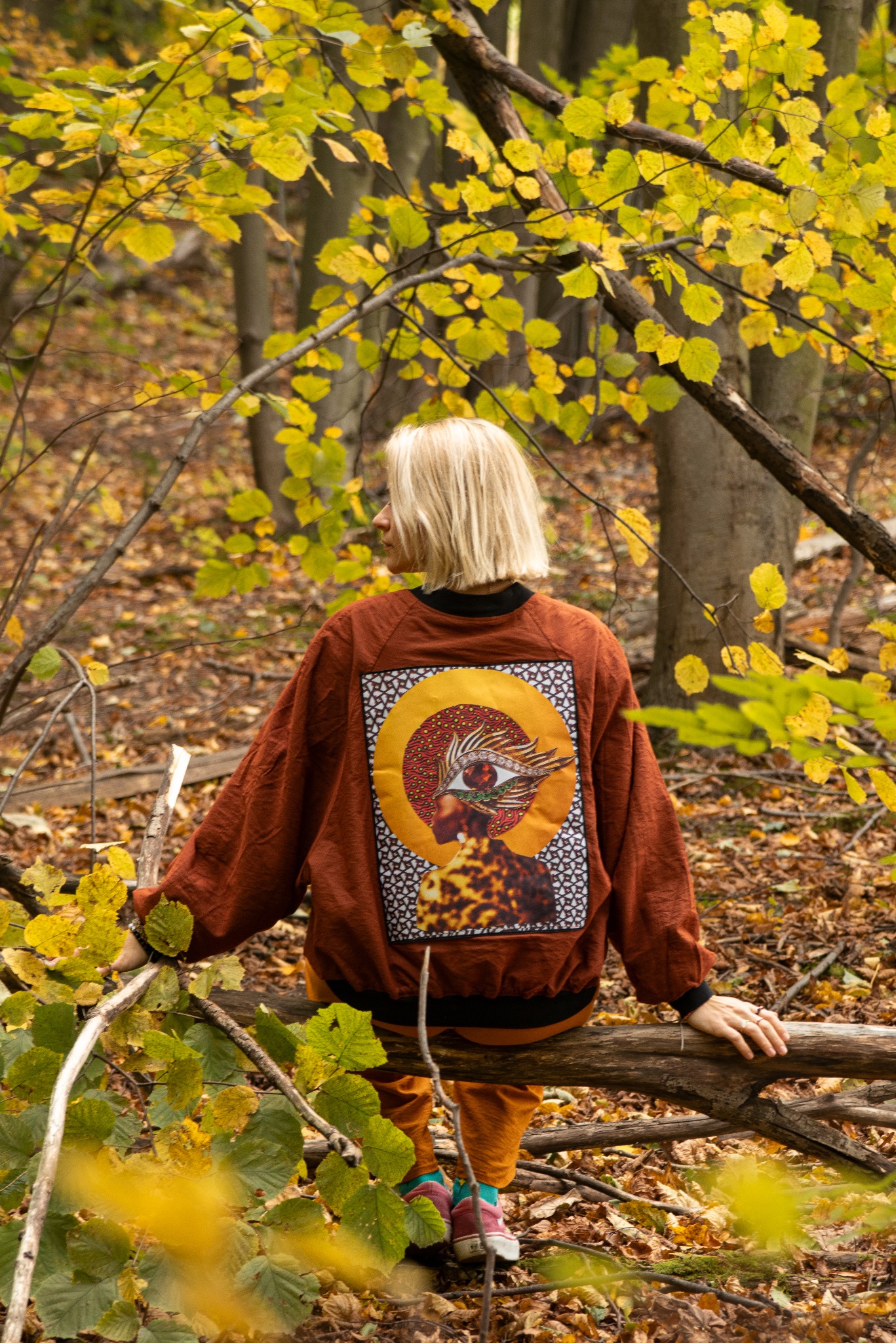 Kobieta z blond włosami siedzi na powalonym drzewie w lesie. Na brązowej bluzie widnieje grafika z okiem i motywami zwierzęcymi. Jesienne liście w tle.