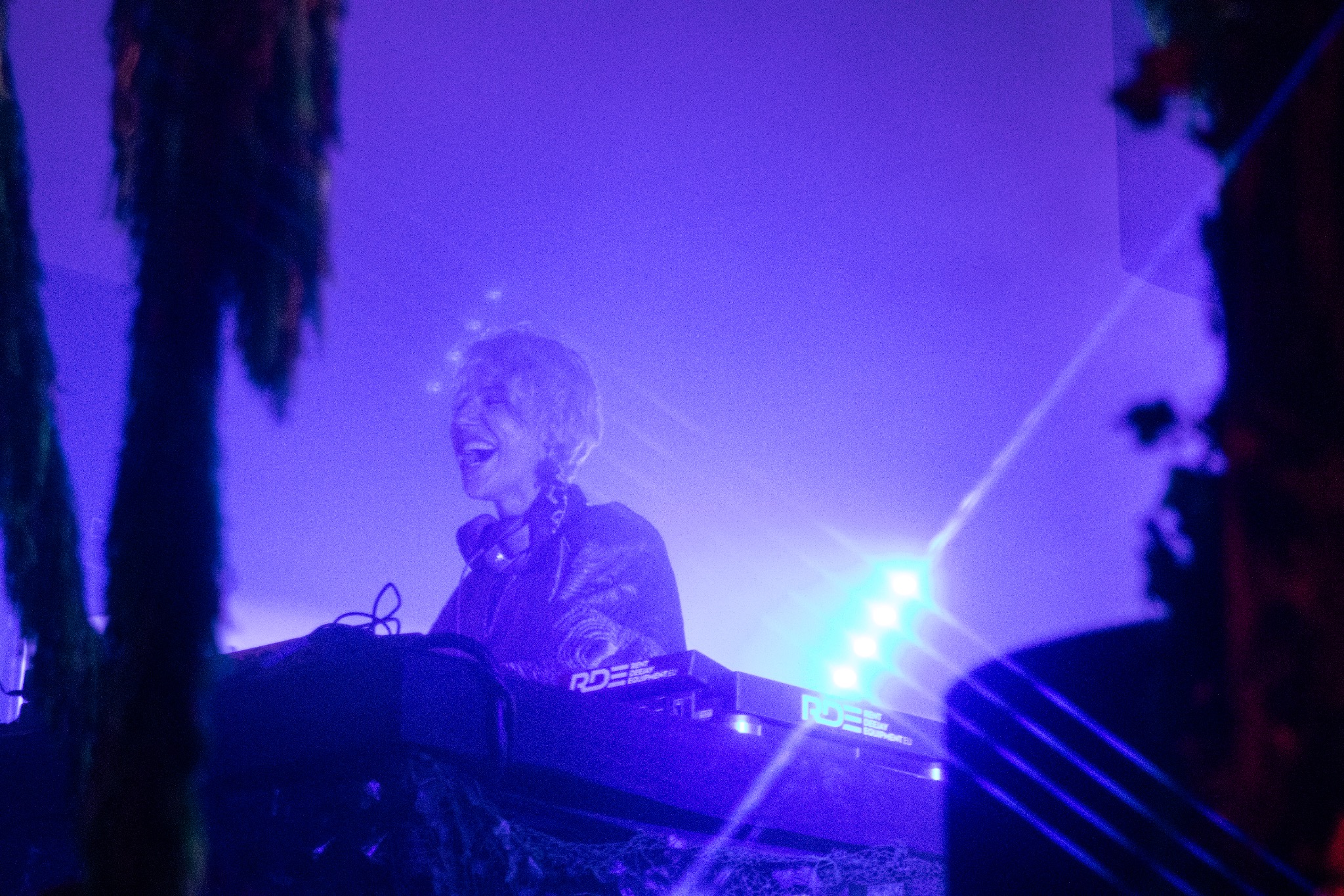 Uśmiechnięta DJ-ka w bluzie ze słuchawkami, grająca na syntezatorze pod fioletowym oświetleniem scenicznym, z elementami dekoracji po bokach kadru.