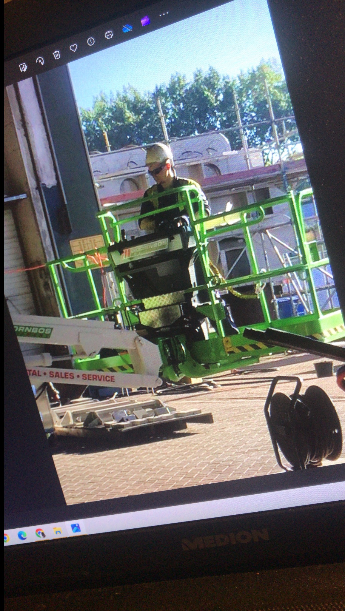 Pracownik w podnośniku koszowym, w okularach ochronnych, wykonuje prace na wysokości przy elewacji budynku. Widoczne rusztowania i elementy konstrukcyjne.