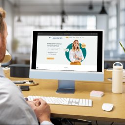 LICHO NIE ŚPI SPÓŁKA Z OGRANICZONĄ ODPOWIEDZIALNOŚCIĄ - Mężczyzna przy biurku ogląda stronę internetową firmy ubezpieczeniowej 'Licho Nie Śpi' na monitorze komputera. Widoczny fragment biura i rośliny doniczkowej.