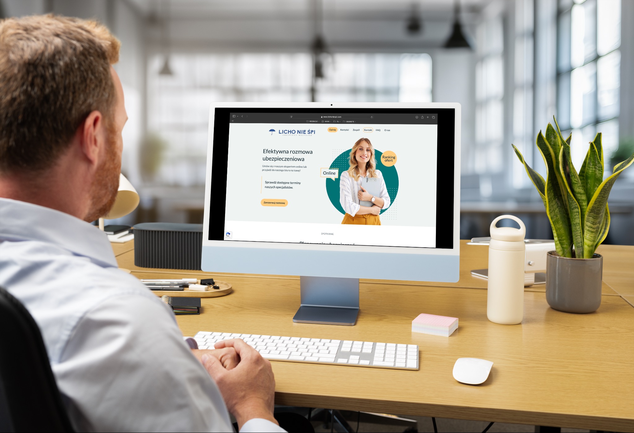 Mężczyzna przy biurku ogląda stronę internetową firmy ubezpieczeniowej 'Licho Nie Śpi' na monitorze komputera. Widoczny fragment biura i rośliny doniczkowej.