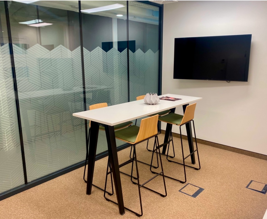 Nowoczesna sala konferencyjna z wysokim stołem, czterema krzesłami barowymi i telewizorem na ścianie. Na stole dekoracja i materiały informacyjne.