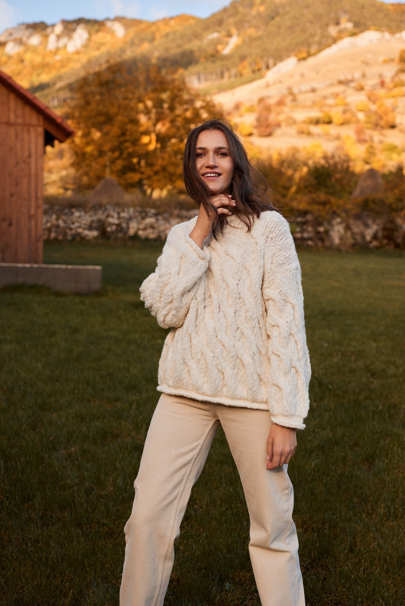 Uśmiechnięta brunetka w kremowym swetrze w warkocze i beżowych spodniach, pozuje na tle jesiennego krajobrazu z drewnianym domkiem i wzgórzami.