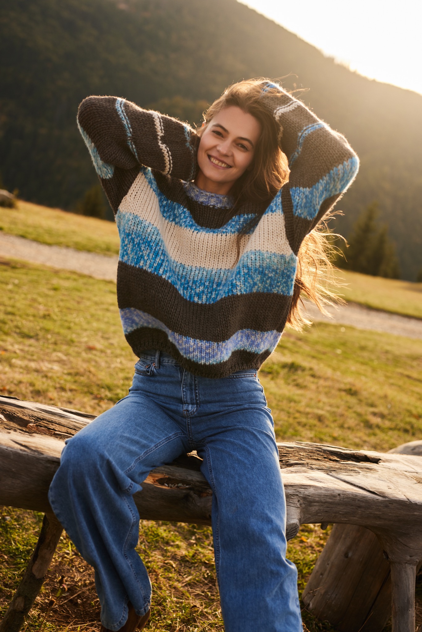 Uśmiechnięta kobieta w szerokich jeansach i grubym, pasiakowym swetrze siedzi na drewnianej ławce na tle górskiego krajobrazu w ciepłym, zachodzącym słońcu.