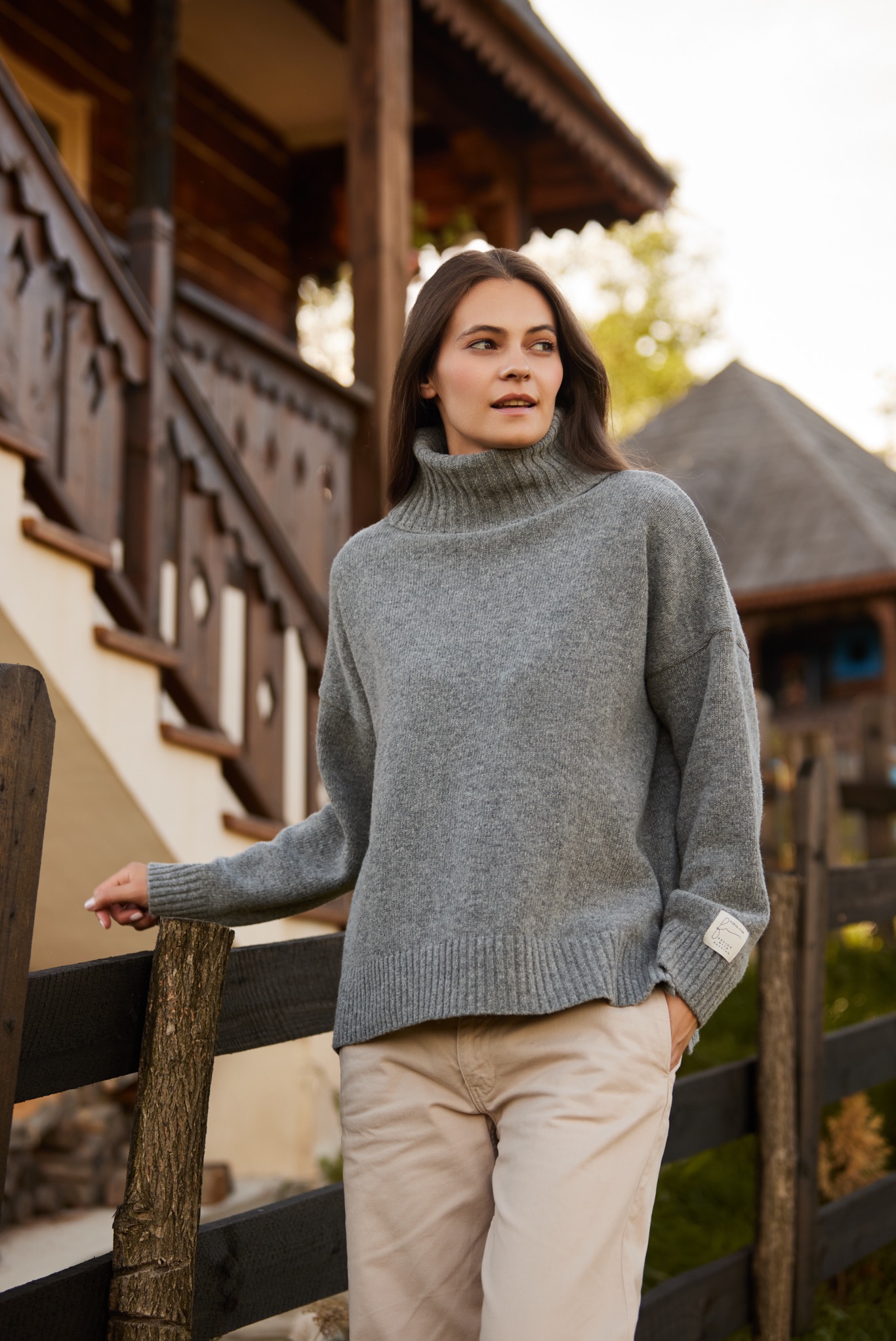 Kobieta w szarym swetrze z golfem i beżowych spodniach, opierająca się o drewniane ogrodzenie na tle drewnianego domu z schodami i charakterystycznym dachem.