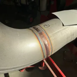 Precyzyjne spawy na rurze ze stali nierdzewnej, tęczowe zabarwienie spoiny, widoczne oznaczenia na materiale, wspornik pod rurą.