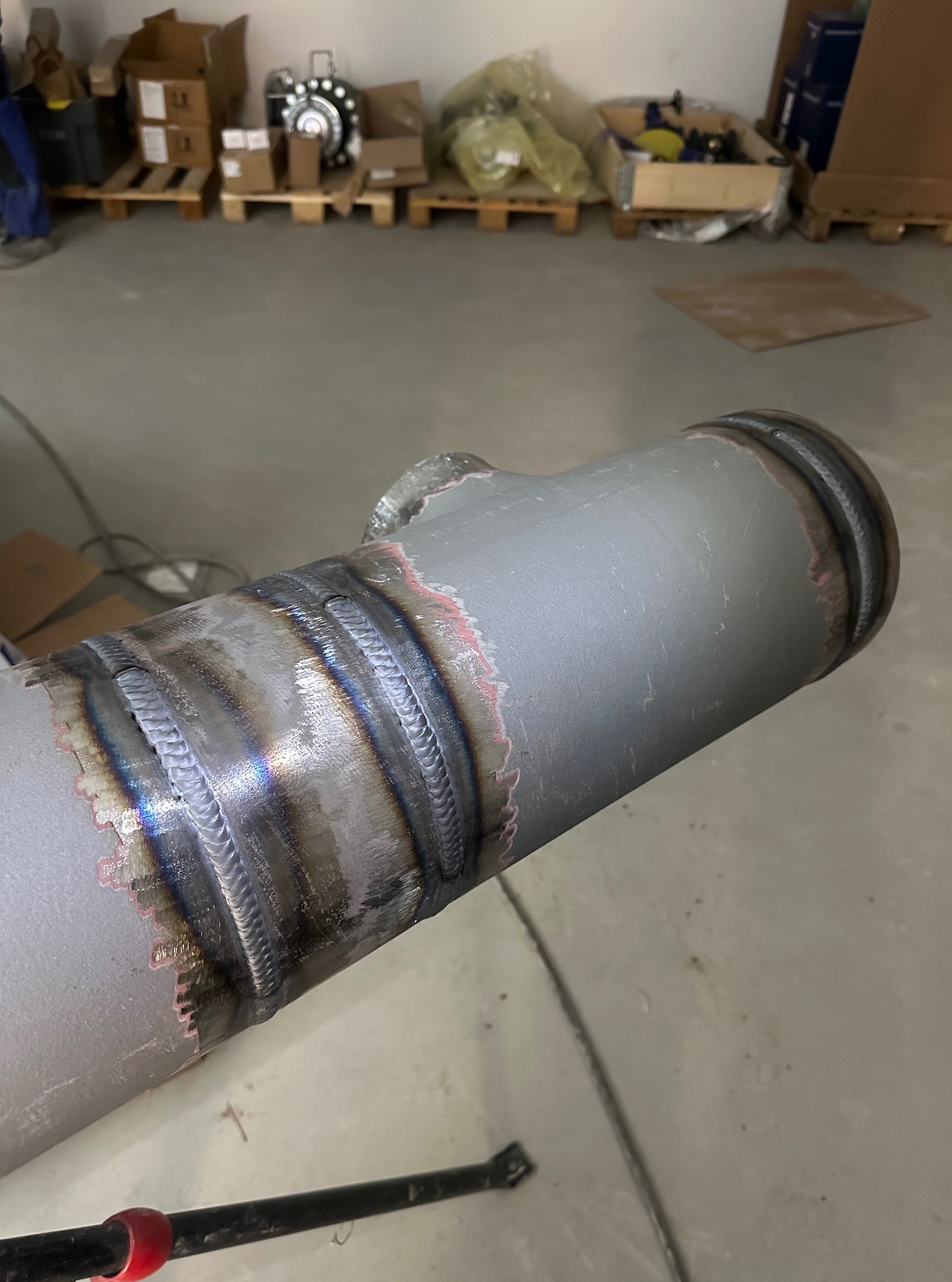 Szara rura z widocznymi, precyzyjnymi spawami, ukazująca solidne łączenia. Tło stanowią kartony i palety w hali.