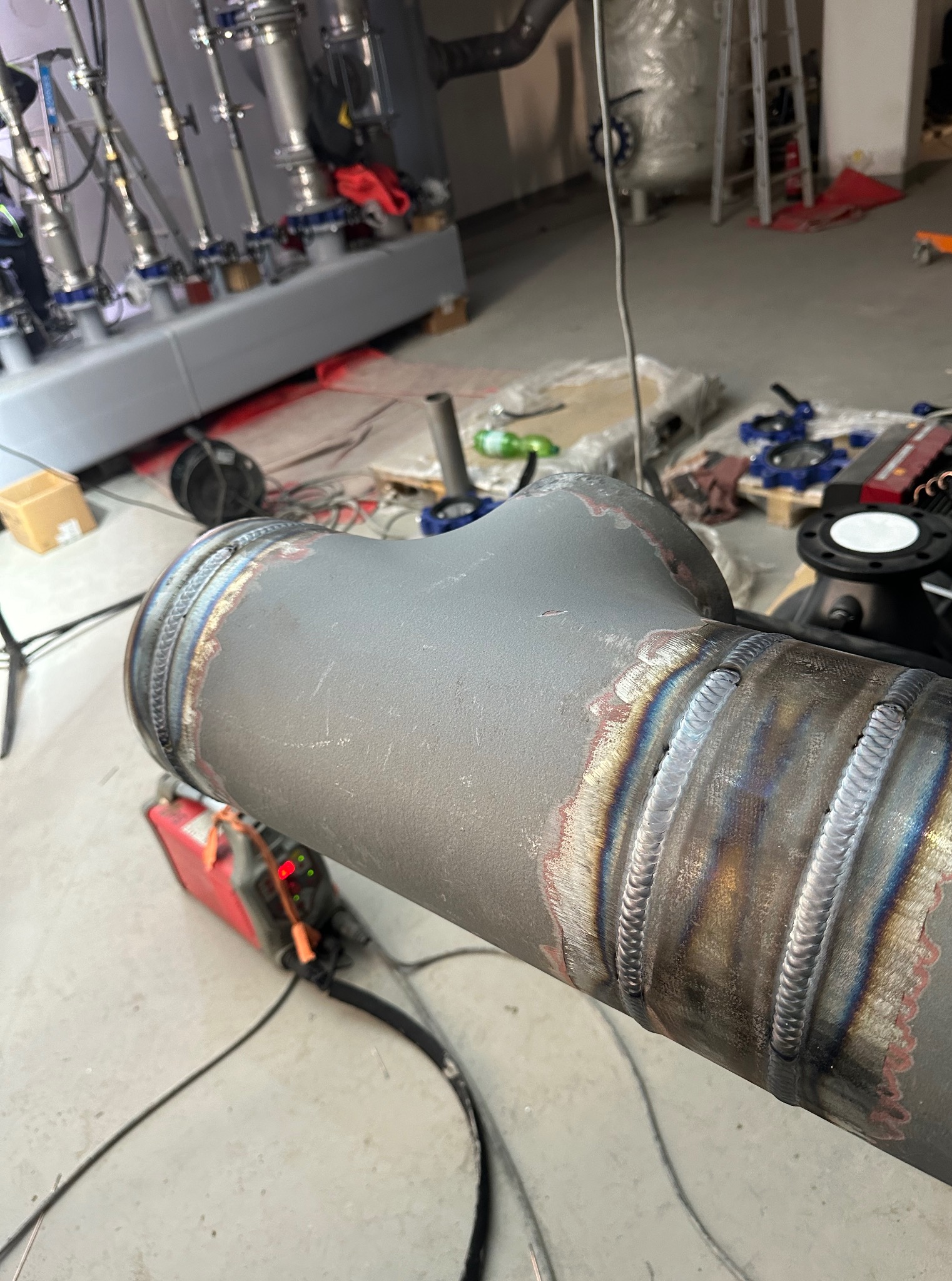 Szlifowany trójnik stalowy z widocznymi spawami, leżący w warsztacie, obok czerwona spawarka i elementy instalacji. Detal spawalniczy z bliska.