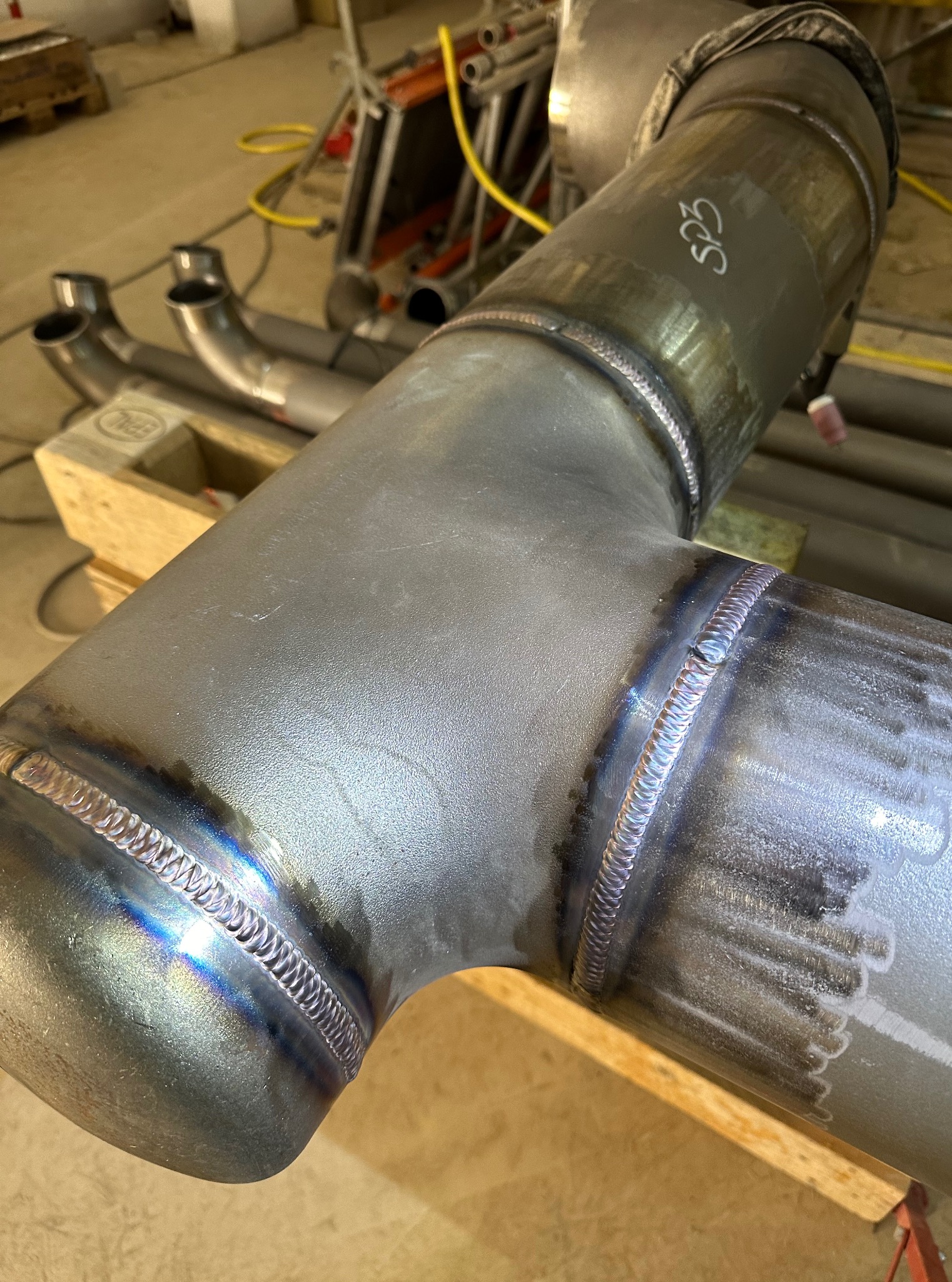 Metalowa rura spawana metodą TIG, z widocznymi spoinami i oznaczeniem 'S23'. W tle elementy warsztatu, w tym inne rury i rusztowanie.