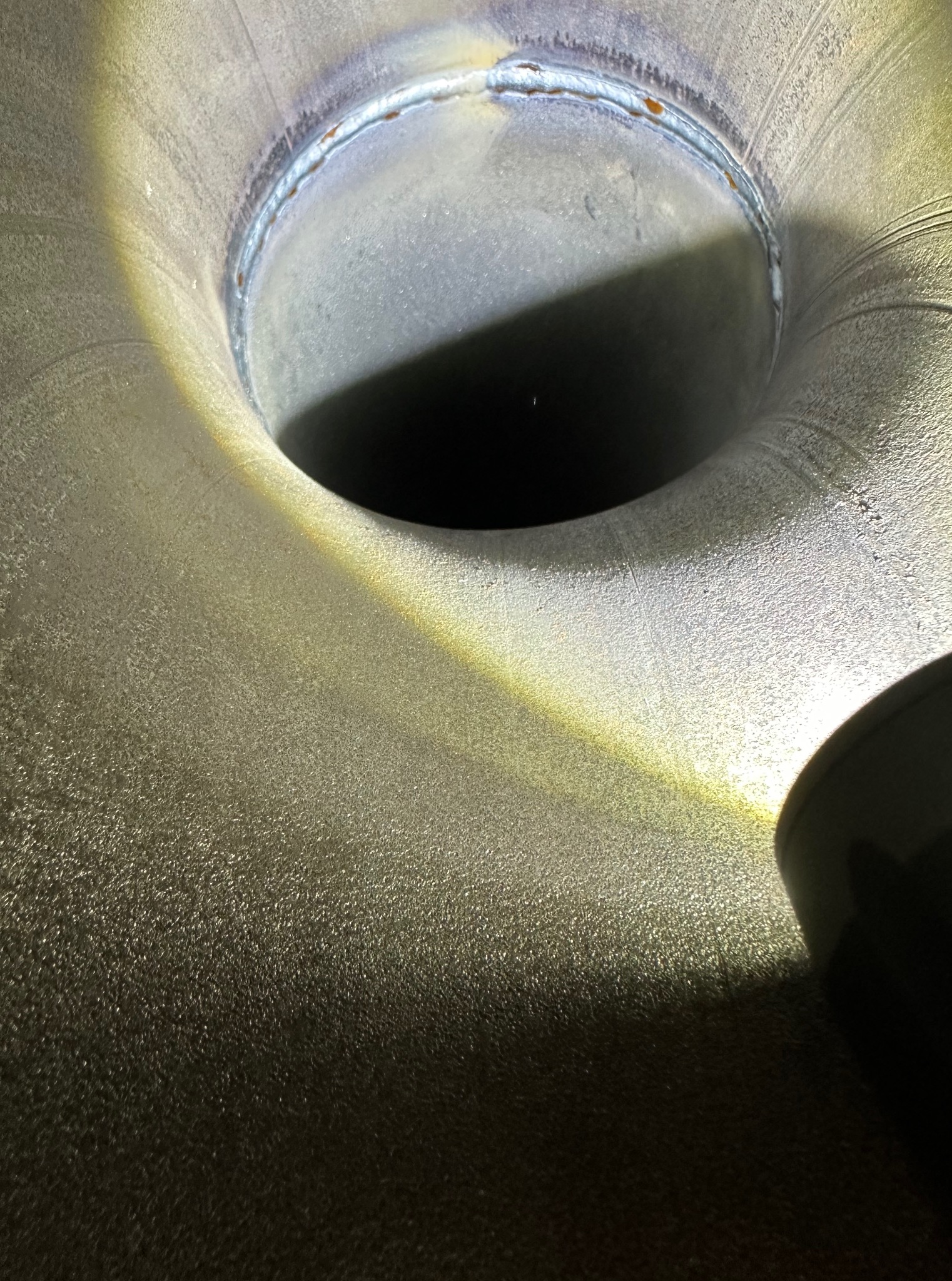 Szlifowana, metalowa rura z widocznym spawem okrężnym, oświetlona punktowo. Widoczna faktura metalu i ślady obróbki.