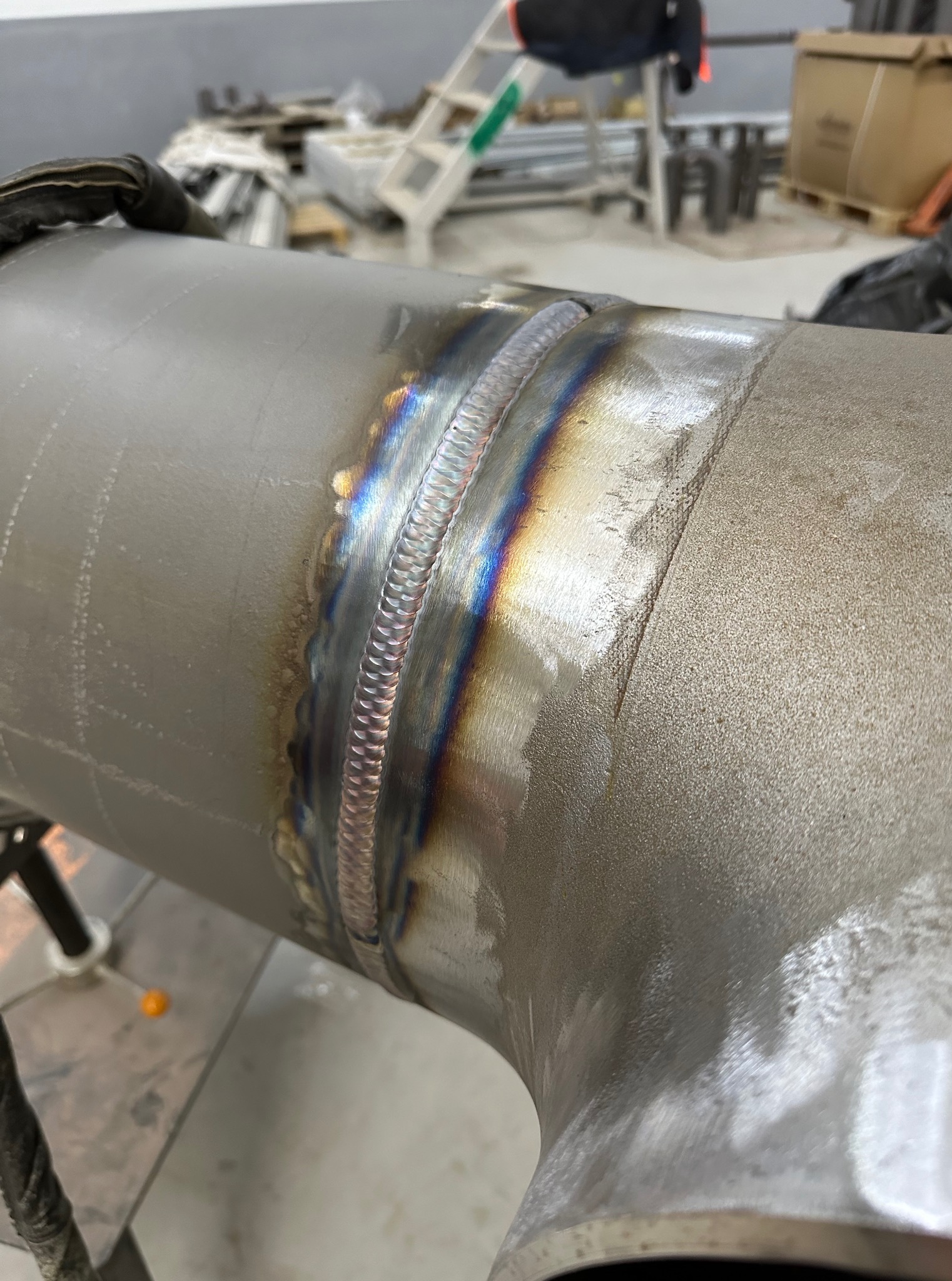 Szlifowany, spawany element rurowy ze stali nierdzewnej z widocznymi śladami spawania TIG i tęczowymi przebarwieniami na spoinie. W tle drabina i kartony.