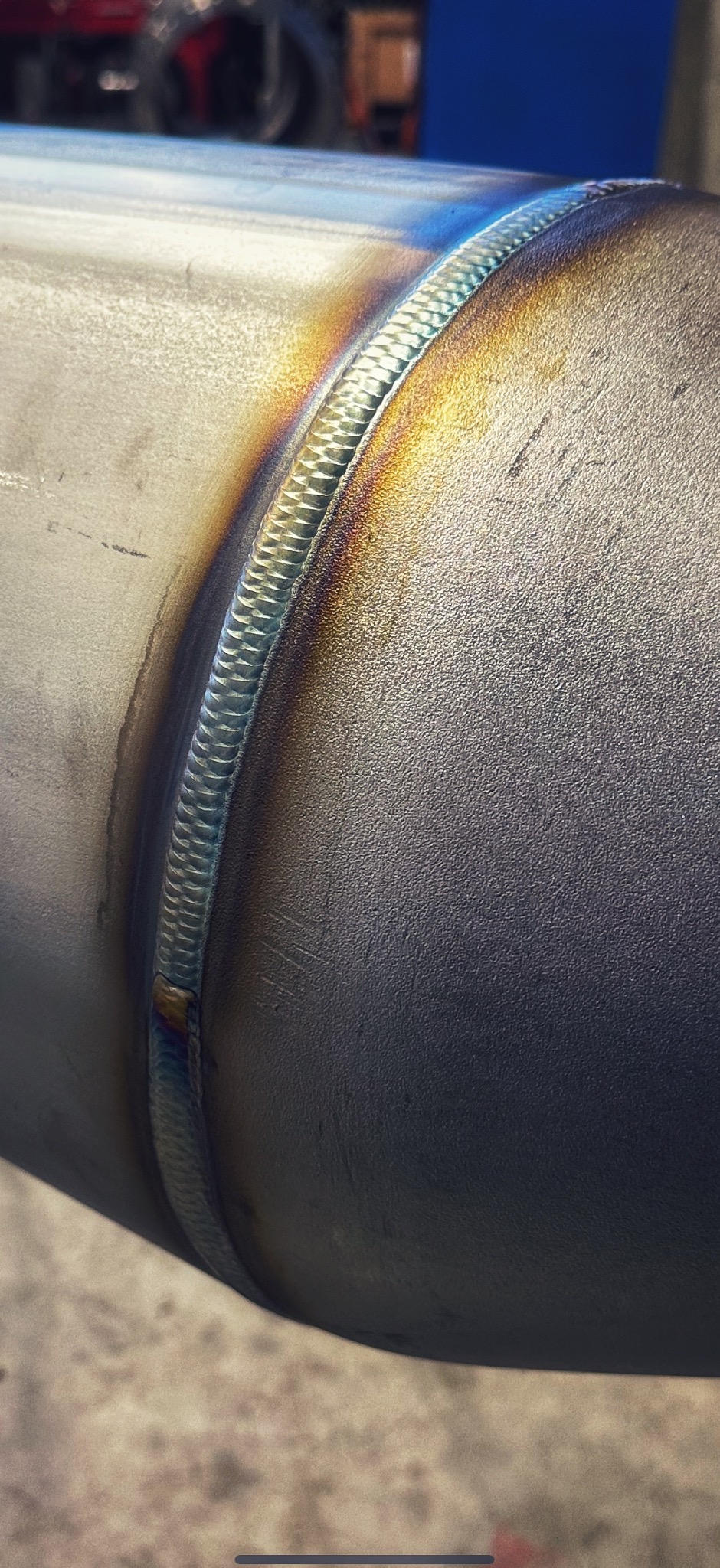 Szczegółowe zbliżenie spawu na metalowej rurze, ukazujące precyzyjne łączenie z widocznymi kolorami hartowania. Widoczne drobne rysy na powierzchni.