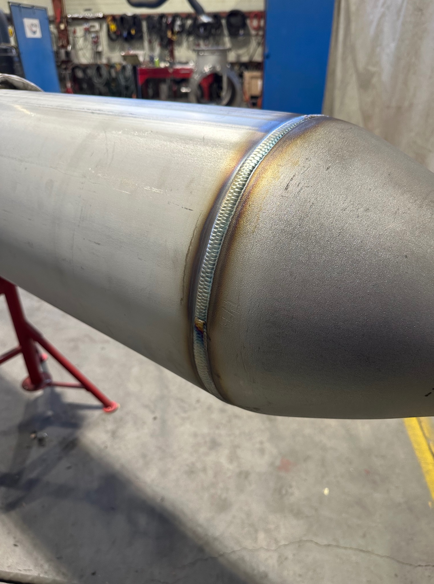 Szlifowany, spawany element ze stali nierdzewnej na czerwonym stojaku. Widoczny spaw na łączeniu stożka i cylindra. Tło warsztatowe.