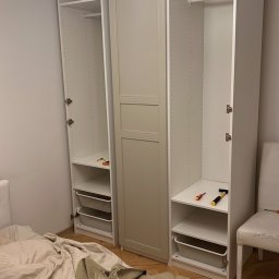 Rafał Marchewka - Montaż białej szafy wnękowej z półkami i koszami w pokoju, widoczne narzędzia, częściowo rozścielone łóżko z pościelą i instrukcja montażu.