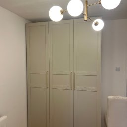 Rafał Marchewka - Trzydrzwiowa szafa w odcieniach beżu z złotymi uchwytami, oświetlona lampą sufitową z kulistymi kloszami, w minimalistycznym wnętrzu.