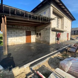 Rafał Marchewka - Nowa, czarna kostka brukowa ułożona na tarasie domu z oświetleniem girlandą. Mężczyzna w czerwonej koszulce przy ścianie. Widoczne narzędzia i worki z materiałami.