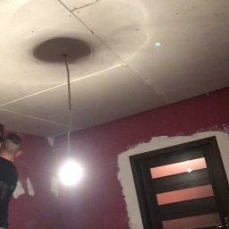 Rafał Marchewka - Montaż płyt karton-gips na suficie w pokoju z bordowymi ścianami. Widoczny pracownik podczas pracy i fragment drzwi. Sufit z okrągłym elementem dekoracyjnym.