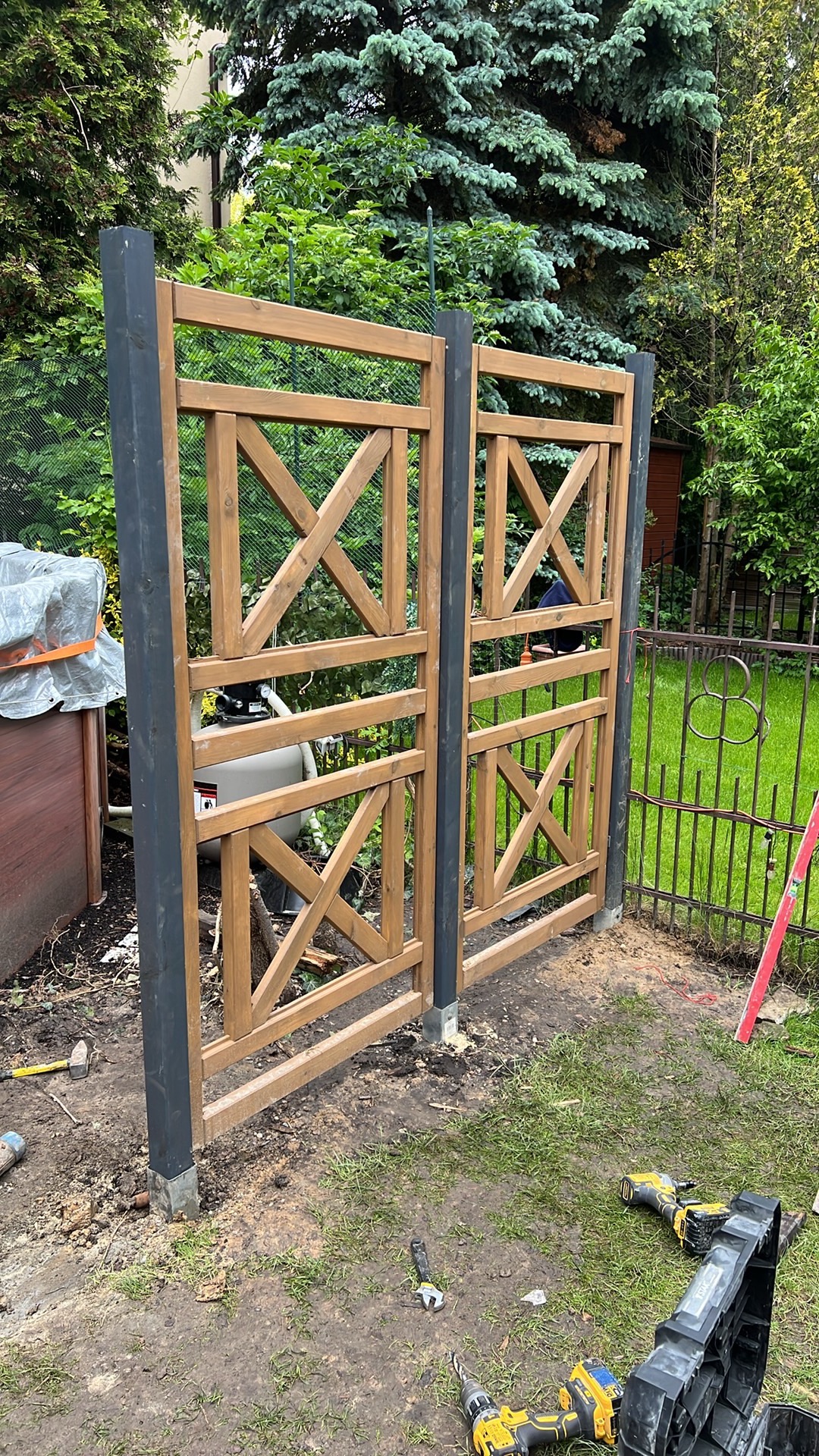 Dwie drewniane kratownice z czarnymi słupkami, tworzące ogrodzenie w ogrodzie. Widoczne narzędzia i elementy montażowe na ziemi. W tle zieleń drzew i krzewów.