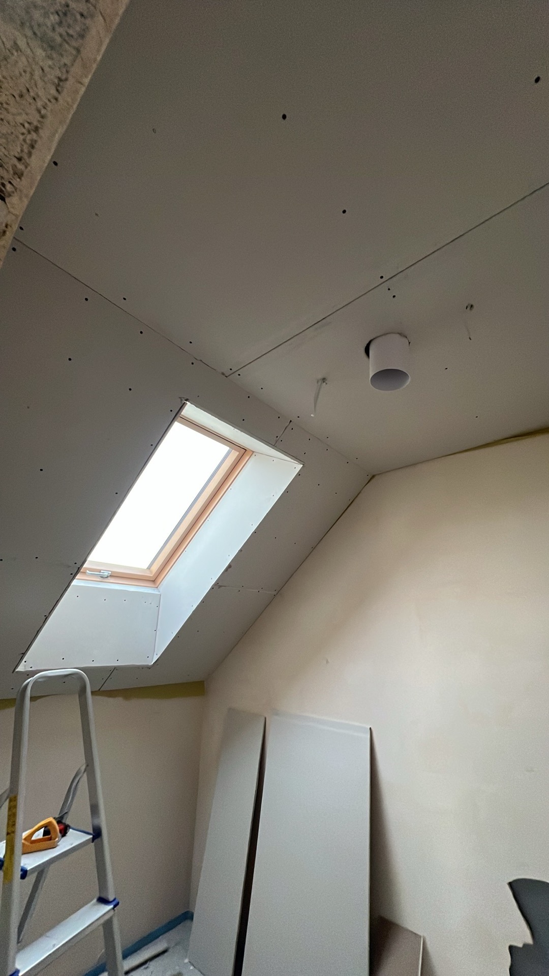 Poddasze z oknem dachowym po montażu płyt kartonowo-gipsowych. Widoczna drabina i przygotowane płyty. Prace wykończeniowe w toku.
