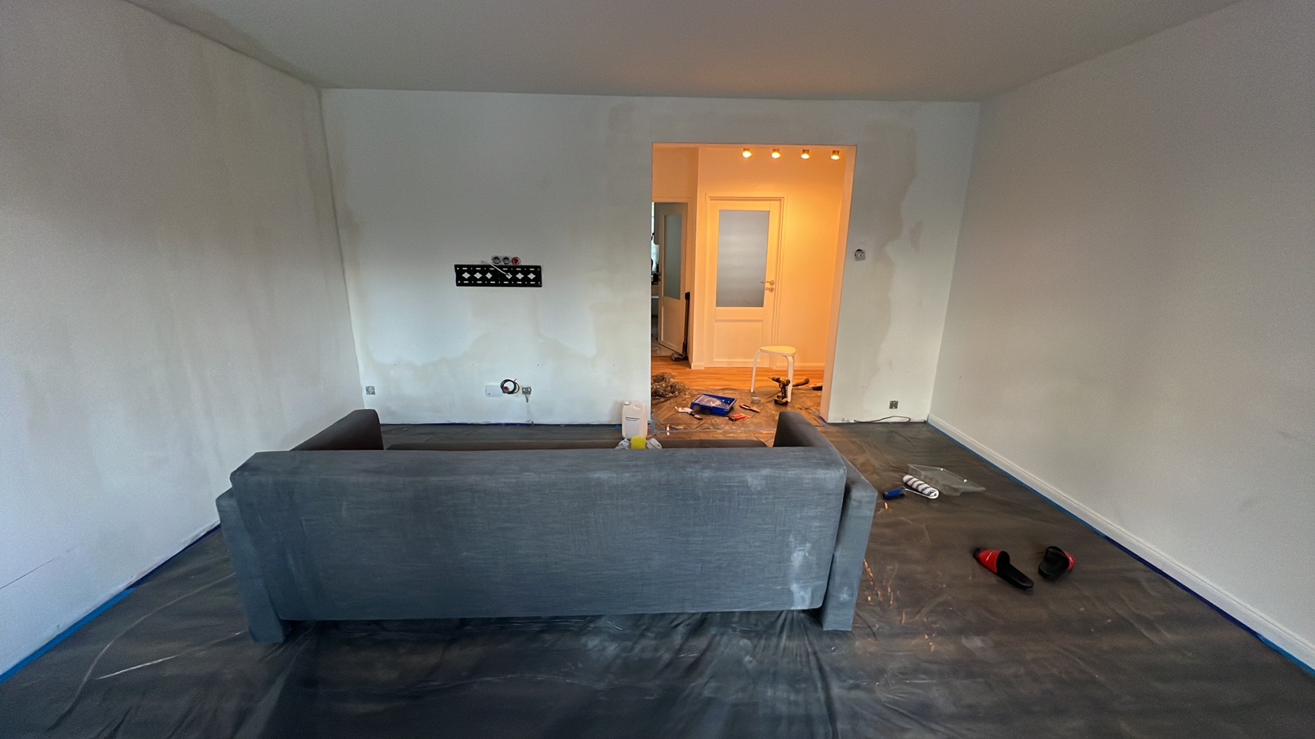 Pokój w trakcie odświeżania: białe ściany, zabezpieczona folią podłoga, widoczny fragment sofy i akcesoria malarskie. W tle przejście do innego pomieszczenia.