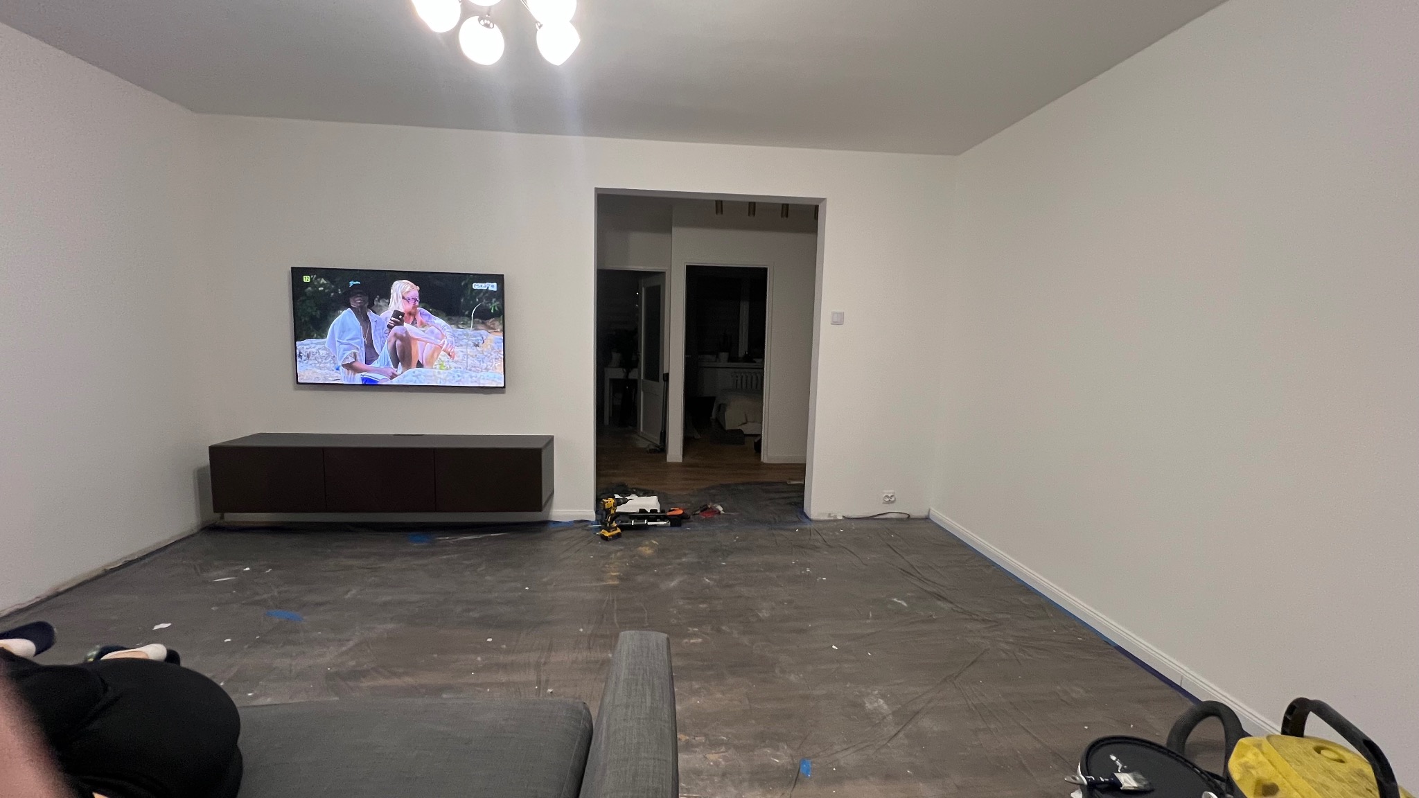 Pokój przygotowany do malowania: zabezpieczona folią podłoga, ściany świeżo pomalowane na biało, widoczny sprzęt malarski i telewizor na ścianie.