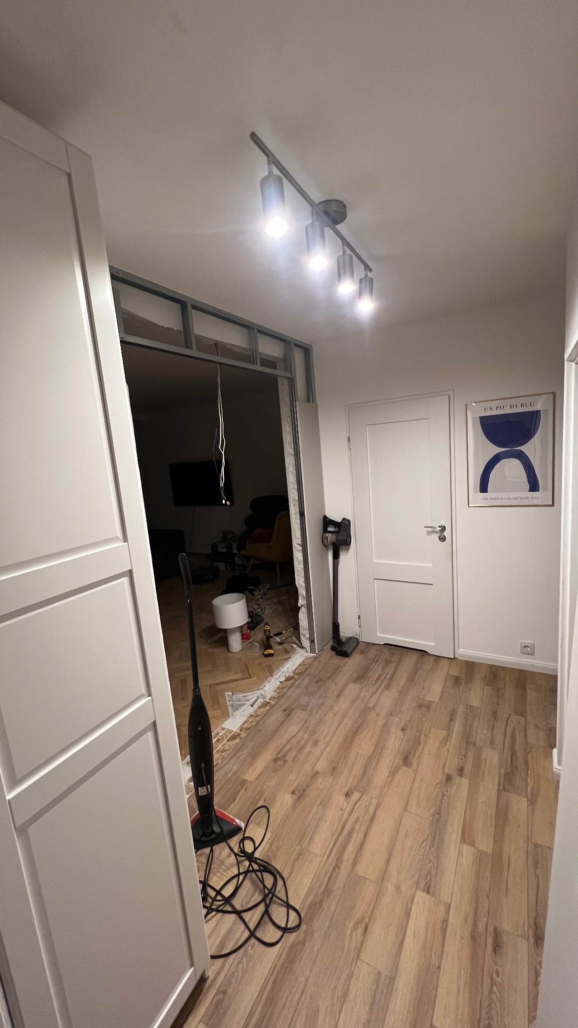 Wnętrze w trakcie remontu: widoczna konstrukcja z profili, podłoga z paneli, narzędzia i sprzęt. Oświetlenie sufitowe, drzwi i plakat na ścianie.