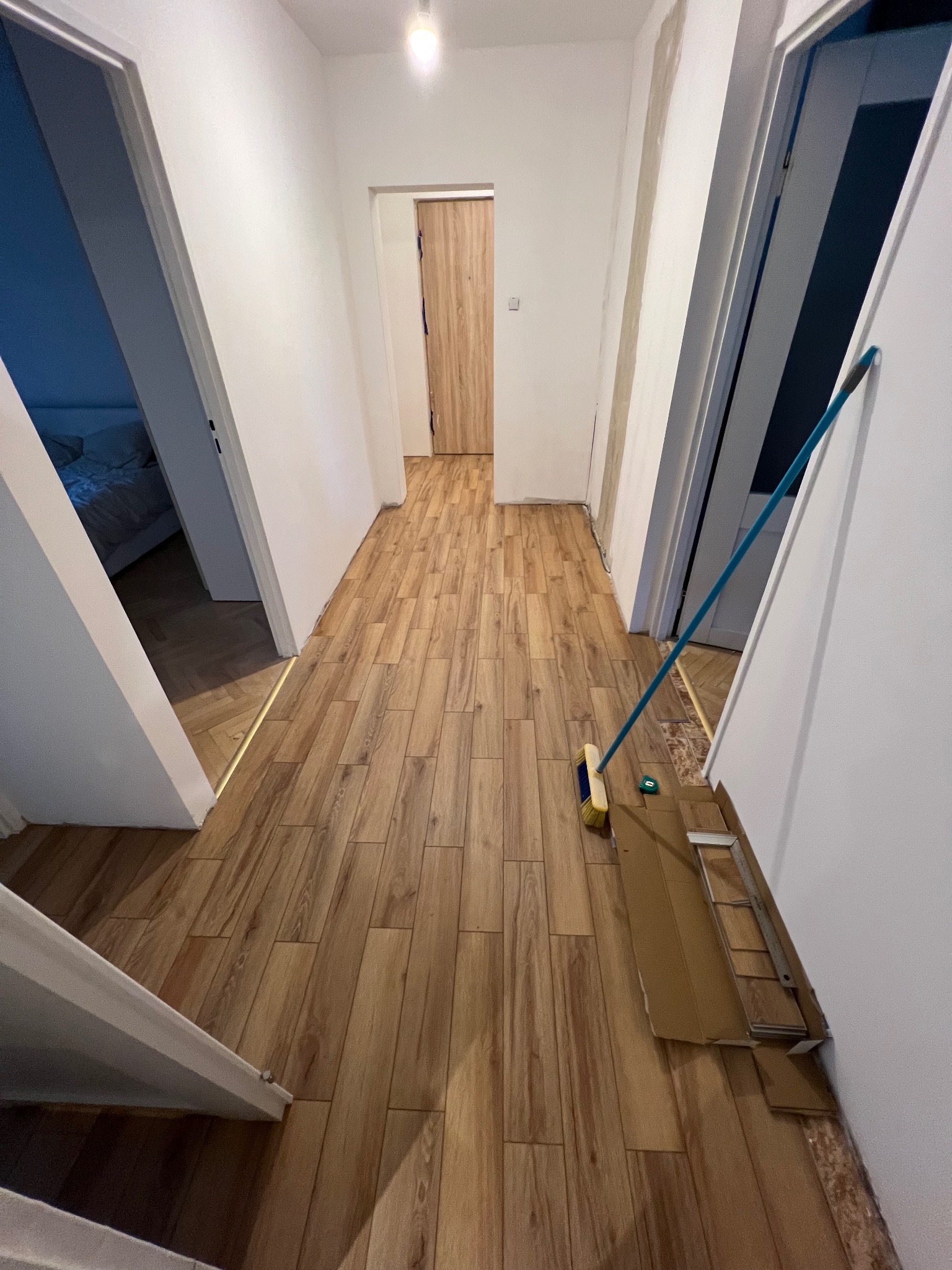 Korytarz po remoncie z nową podłogą imitującą drewno. Widoczne niedoróbki przy drzwiach i listwach. Na podłodze leżą panele i stoi zmiotka.