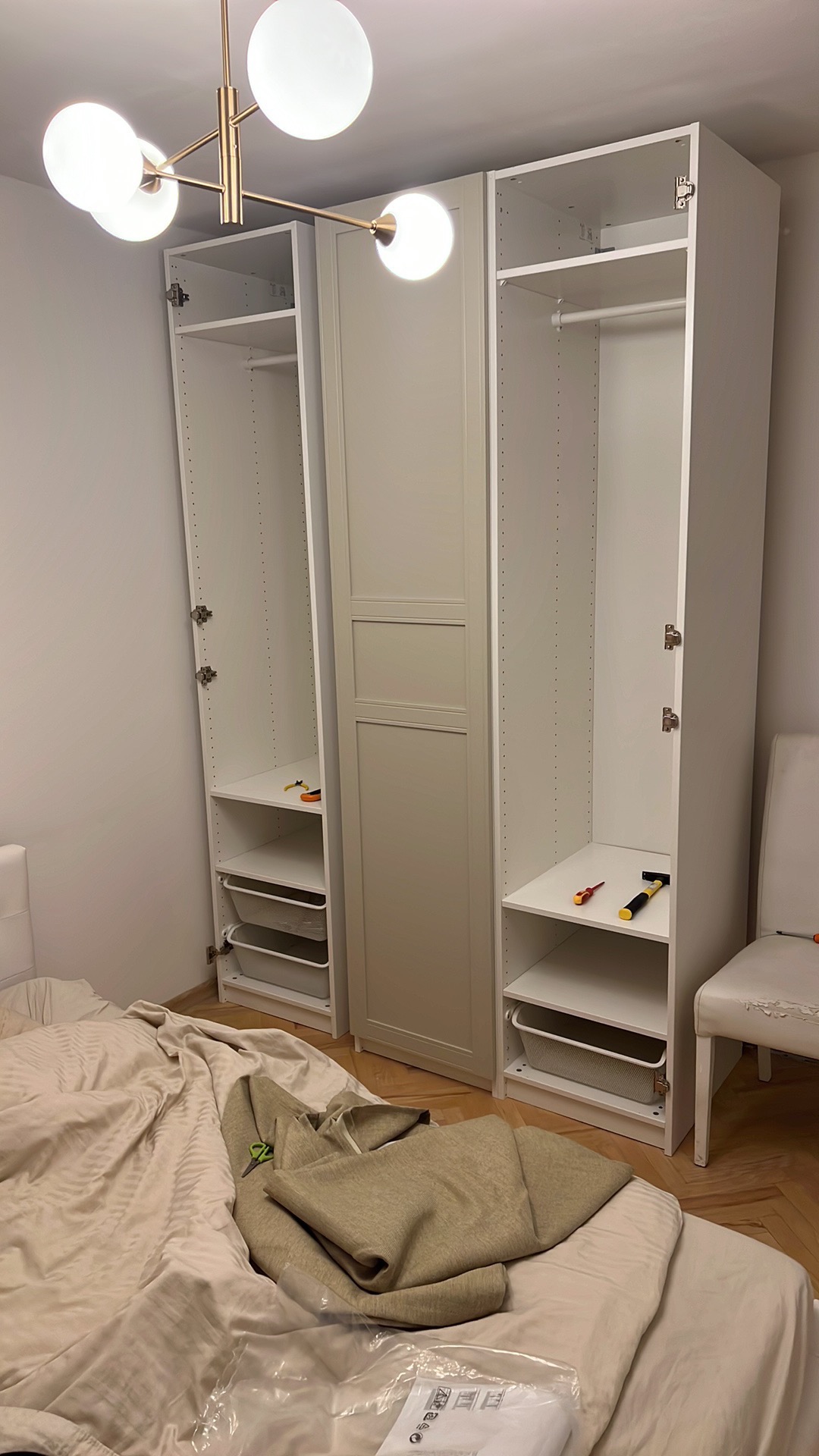 Montaż białej szafy wnękowej z półkami i koszami w pokoju, widoczne narzędzia, częściowo rozścielone łóżko z pościelą i instrukcja montażu.